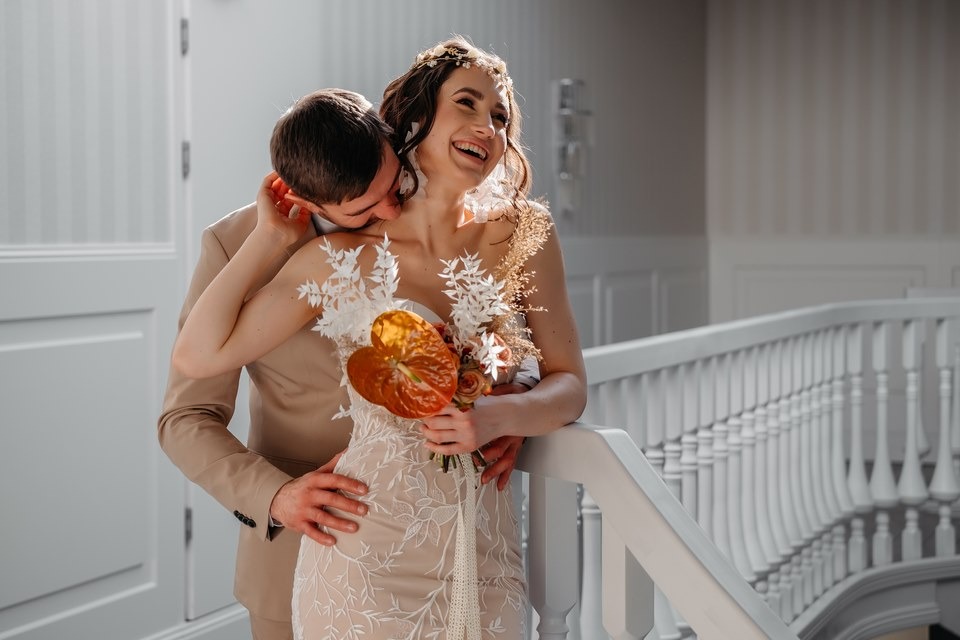 Uśmiechnięta panna młoda w sukni z koronkowymi aplikacjami trzyma bukiet z anturium, oblubieniec całuje ją w szyję na tle białej balustrady.