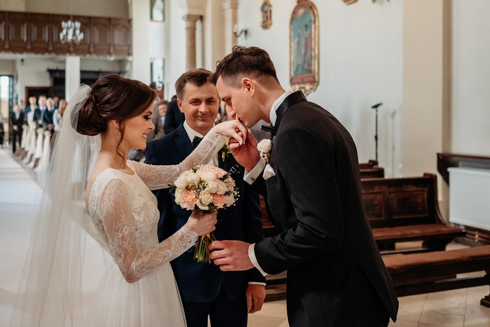 Elegancki pocałunek dłoni panny młodej przez pana młodego w kościele, z uśmiechniętym mężczyzną w tle. Bukiet w rękach panny młodej, ławki i kolumny w tle.