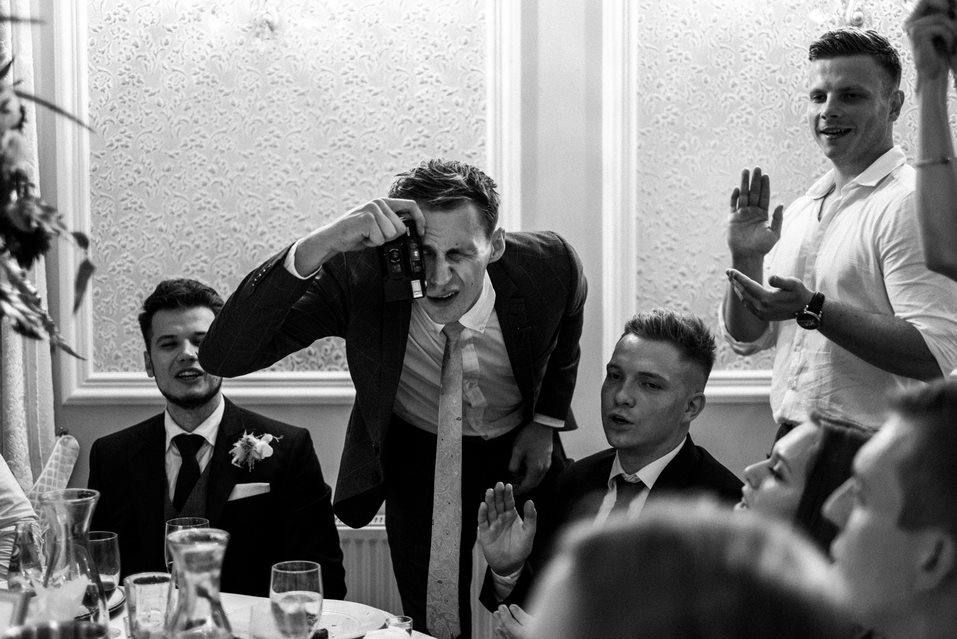 Czarno-białe zdjęcie fotografa z aparatem podczas uroczystości, robiącego zdjęcie gościom siedzącym przy stole. W tle widoczny wzorzysty ornament na ścianie.