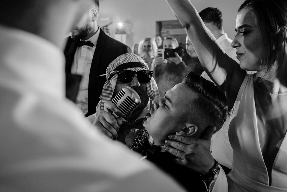 Czarno-białe zdjęcie z imprezy: mężczyzna w kefii trzyma mikrofon przy ustach drugiego mężczyzny, w tle goście i kobieta z podniesioną ręką. Nietypowe ujęcie z zabawy.