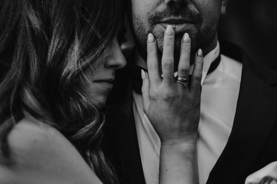 Czarno-białe zbliżenie pary: kobieta z falowanymi włosami opiera się o mężczyznę w smokingu, dłoń z pierścionkiem na jego twarzy. Subtelne światło, intymna atmosfera.