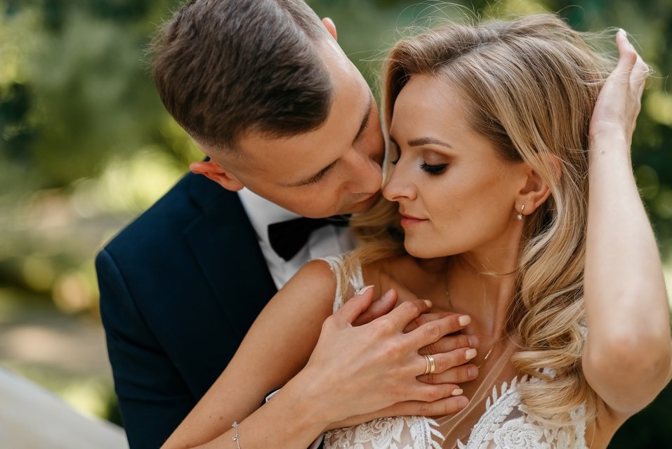 Elegancka para młoda w zbliżeniu: on w granatowym smokingu, ona w koronkowej sukni, delikatny pocałunek na tle zieleni. Romantyczna sesja w plenerze, złote obrączki.