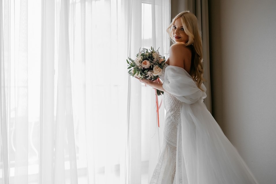 Panna młoda w koronkowej sukni, z bukietem, pozuje przy oknie z białymi zasłonami. Delikatne światło podkreśla detale sukni i makijaż. Uśmiech skierowany w stronę obiektywu.