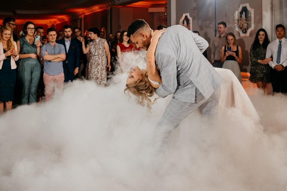 Para młoda tańczy pierwszy taniec w chmurze dymu na sali weselnej, goście obserwują. Pan młody w szarym garniturze w kratę, panna młoda w białej sukni.