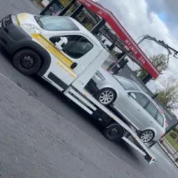 Laweta z srebrnym kombi Audi na platformie, na tle myjni bezdotykowej i pochmurnego nieba. Żółte logo firmy na białym samochodzie pomocy drogowej.