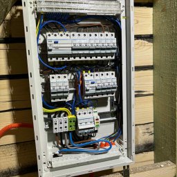 Michał Dudek - Rozdzielnica elektryczna Hager z bezpiecznikami, zamontowana na drewnianej ścianie. Widoczne przewody, szyny DIN i elementy zabezpieczające. Instalacja w trakcie realizacji.