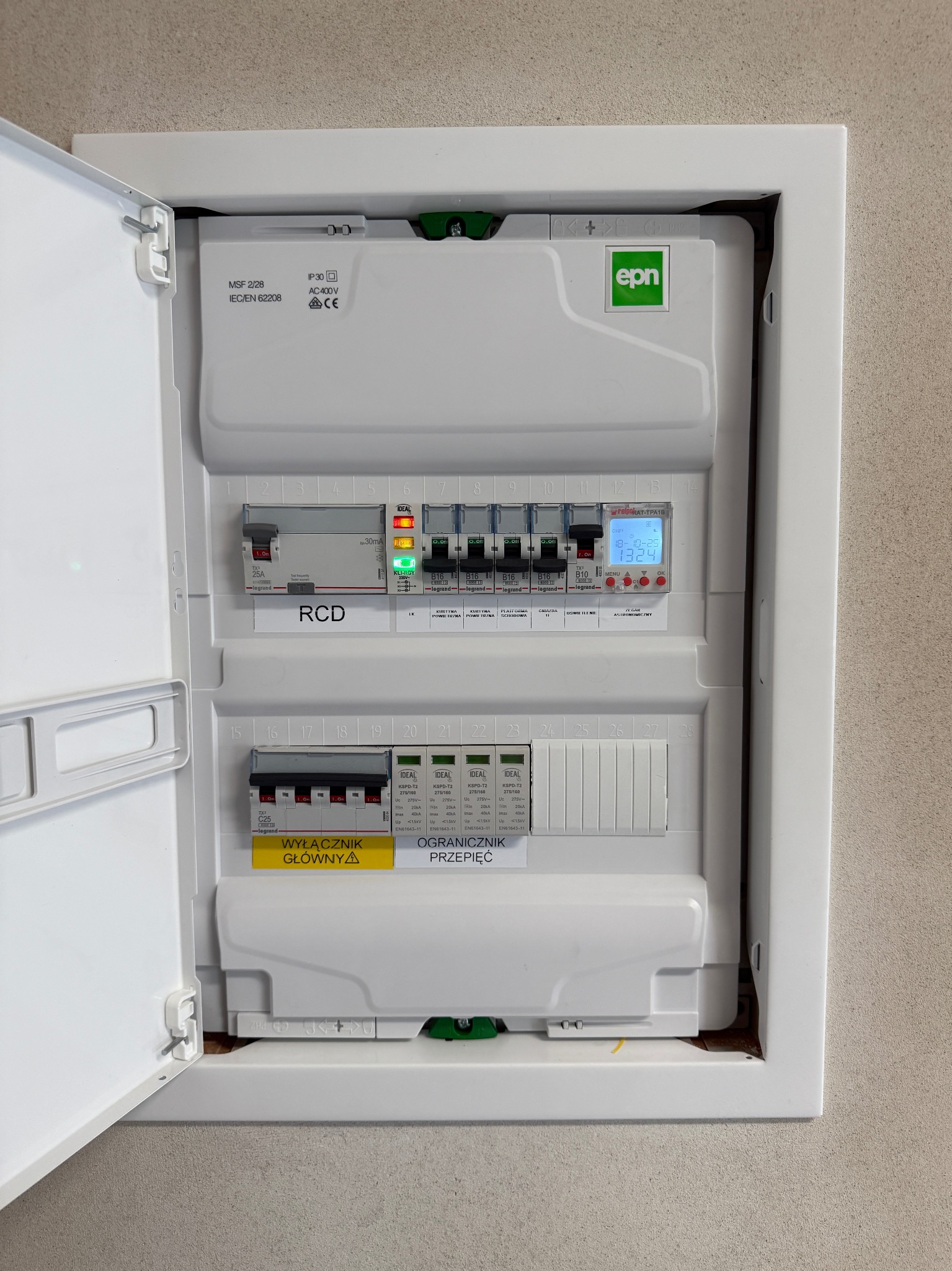 Otwarta skrzynka elektryczna z bezpiecznikami, wyłącznikiem głównym i ogranicznikiem przepięć. Widoczne oznaczenia i parametry urządzeń. Biała obudowa.