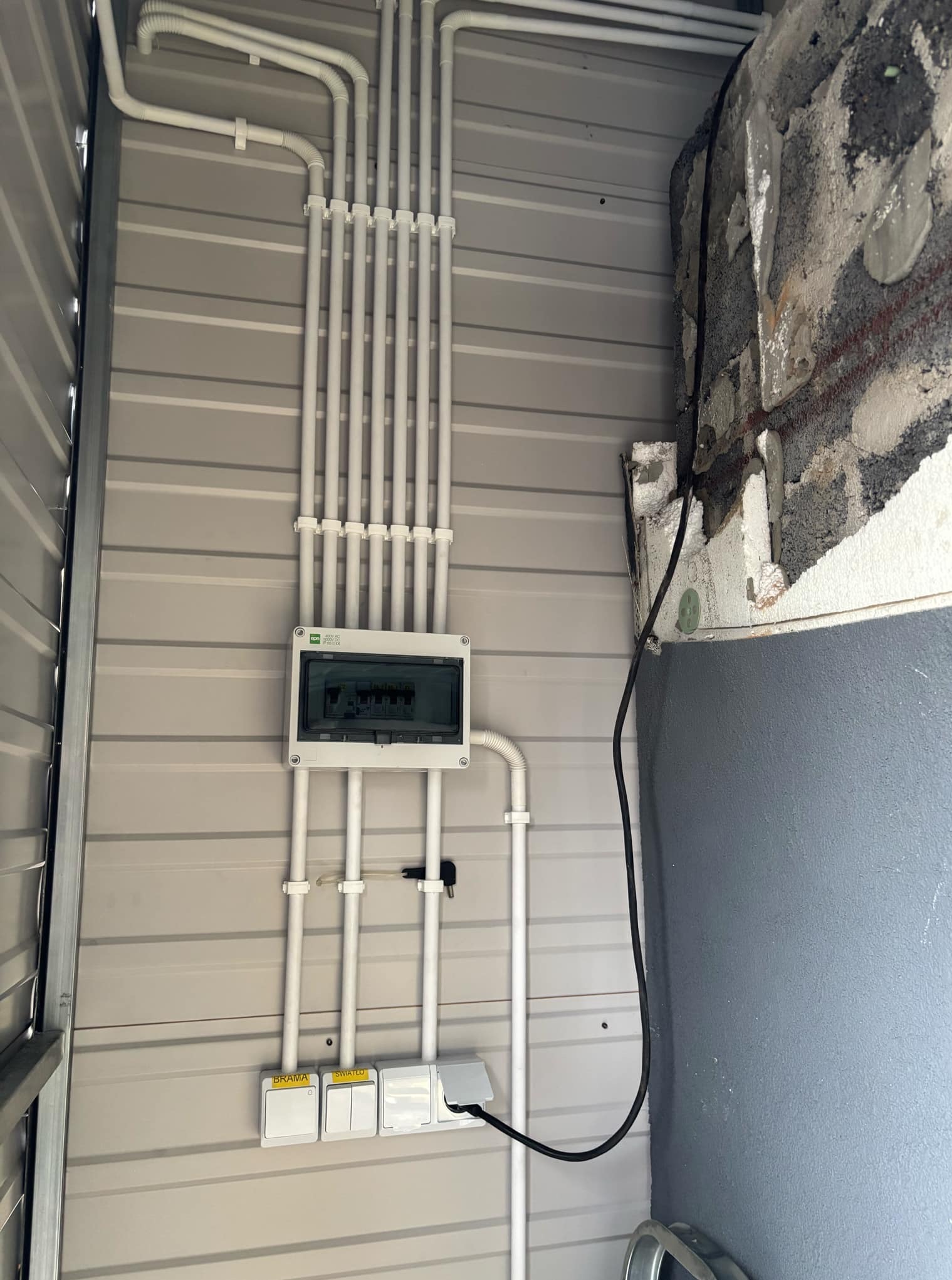 Szczegółowo poprowadzone, białe rury instalacji elektrycznej na metalowej ścianie, widoczna skrzynka z bezpiecznikami i włączniki. Częściowo otynkowana ściana obok.