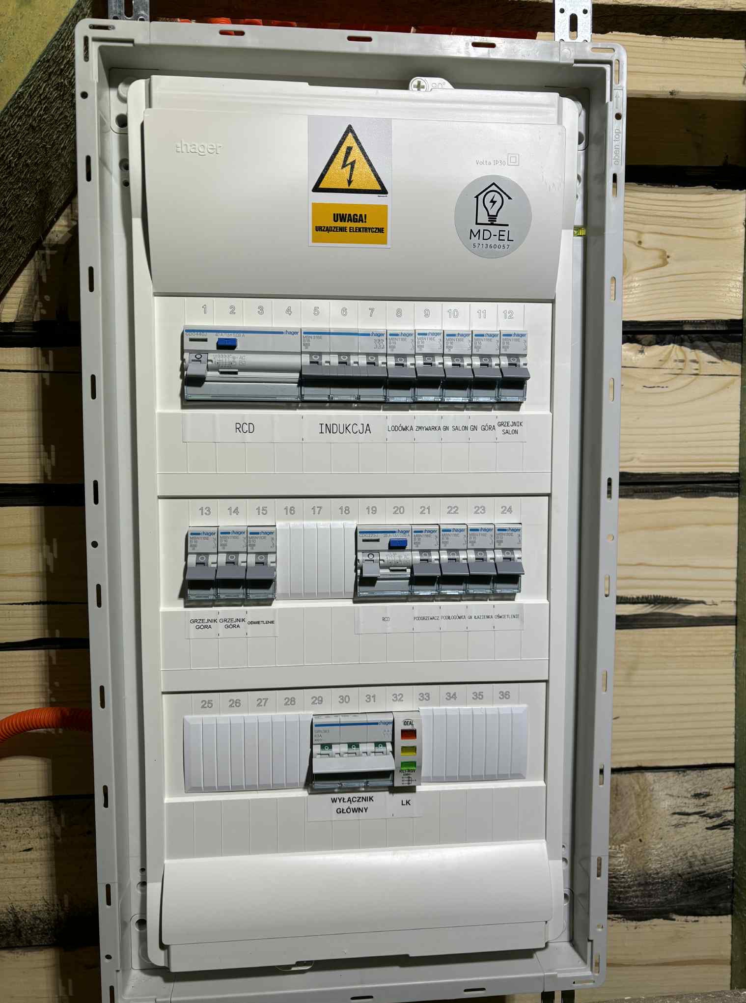 Tablica elektryczna Hager w białej obudowie, z widocznymi bezpiecznikami i wyłącznikami, oznaczona naklejką 'MD-EL'. Zamontowana na ścianie z drewnianych desek.
