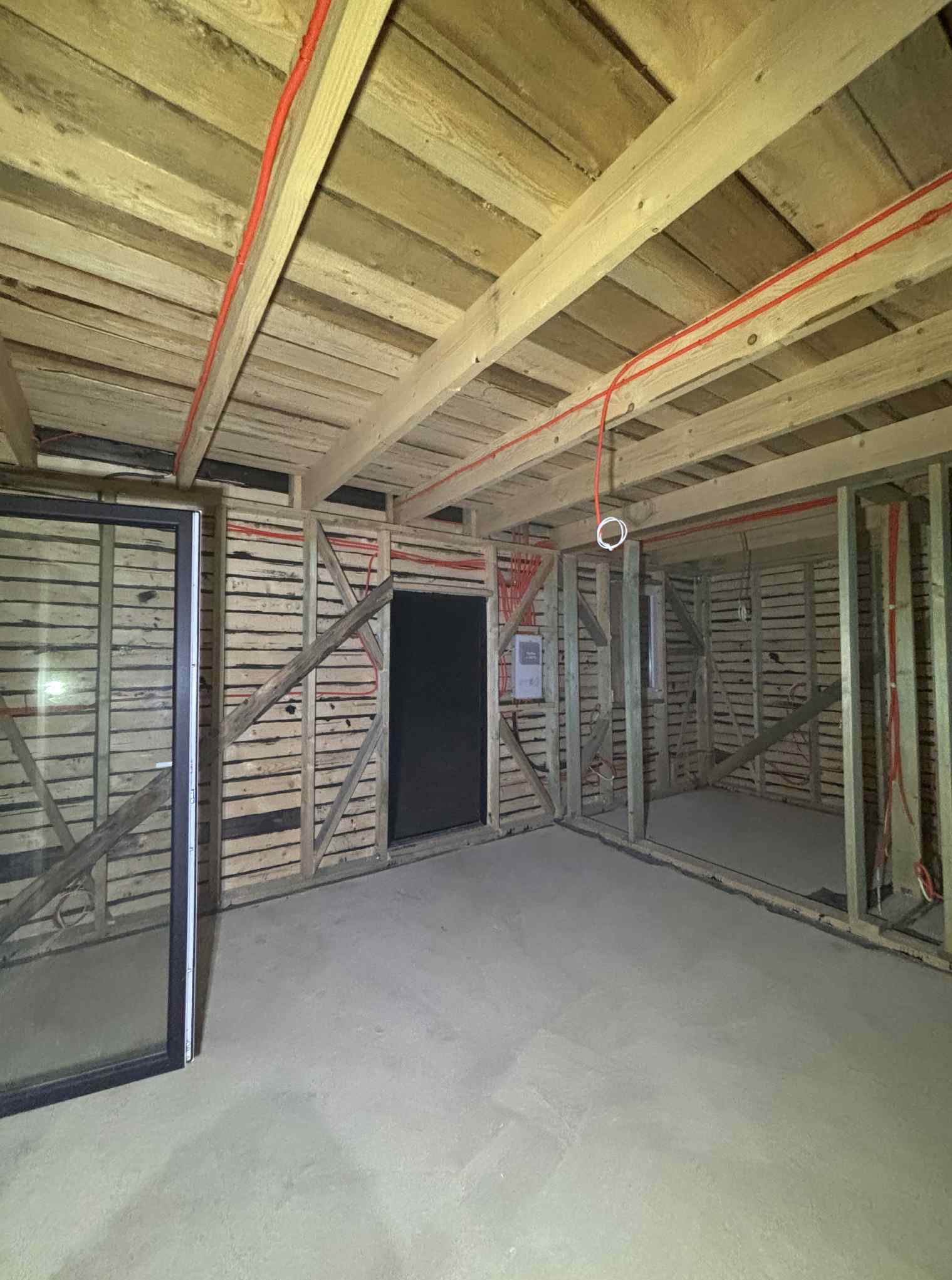Surowy stan wnętrza z drewnianą konstrukcją ścian i stropu, widoczne czerwone przewody elektryczne rozprowadzone po suficie. W tle otwór drzwiowy i okno.
