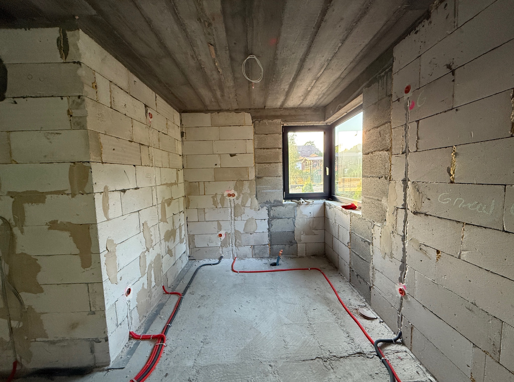 Surowy stan wnętrza z instalacją elektryczną w bruzdach ścian i podłodze. Widoczne kable w czerwonych peszlach, puszki elektryczne. Okno z widokiem na zewnątrz.