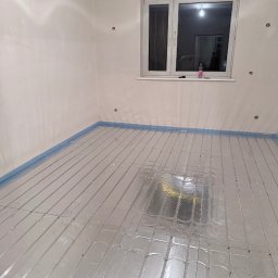 MATEUSZ BEKASIAK MB TECHNIK - Ułożona instalacja ogrzewania podłogowego w pokoju z oknem, zabezpieczona niebieską folią przy ścianach. Widoczne otwory na gniazdka elektryczne.