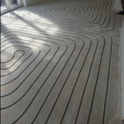 MATEUSZ BEKASIAK MB TECHNIK - Ułożone rury ogrzewania podłogowego w surowym pomieszczeniu, widoczne złącza i fragment okna. Instalacja przed wylaniem posadzki, w jasnym świetle.