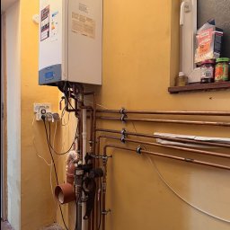 MATEUSZ BEKASIAK MB TECHNIK - Biały piec gazowy Ariston ONE z miedzianą instalacją CO na żółtej ścianie, widok z boku. Na parapecie okna pudełko popcornu i słoiki. Widoczne gniazdka elektryczne.