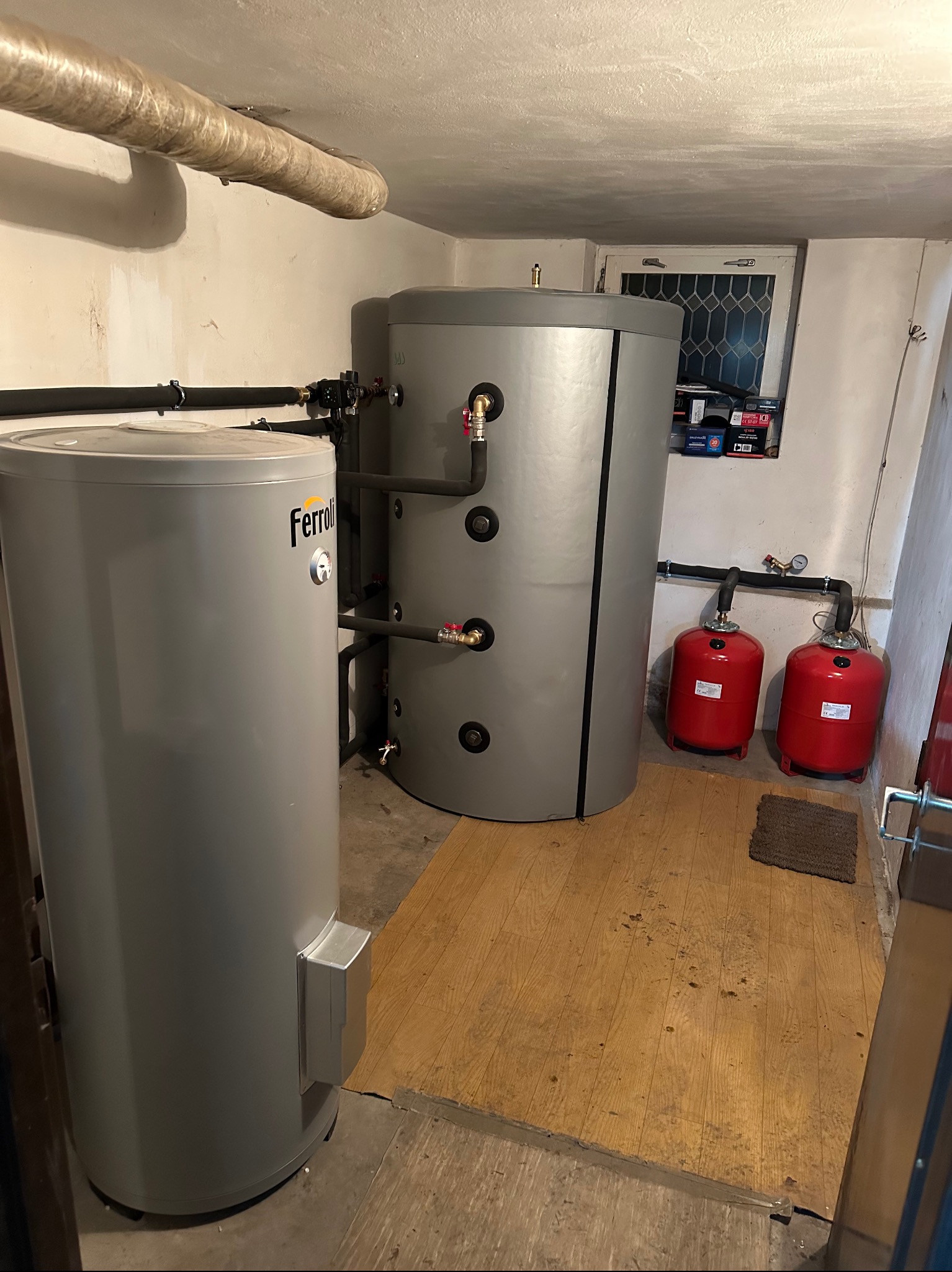 Szare zbiorniki akumulacyjne Ferroli w piwnicy, z rurami i czerwonymi naczyniami wzbiorczymi. Widoczne okno i instalacje na ścianach. Ujęcie z bliska.