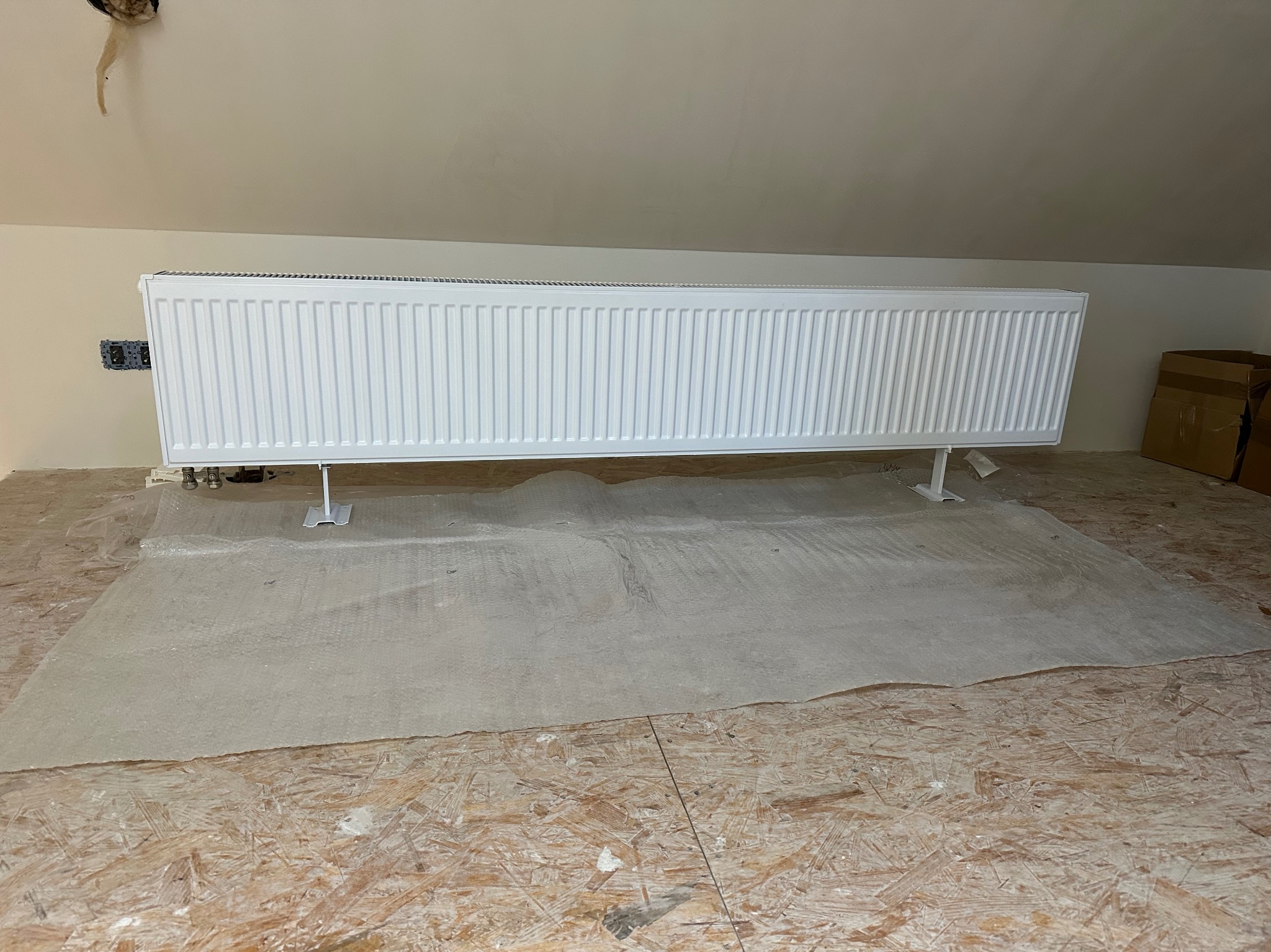 Biały, poziomy grzejnik panelowy na metalowych nóżkach, zamontowany na ścianie w pokoju na poddaszu. Podłoga zabezpieczona folią ochronną.