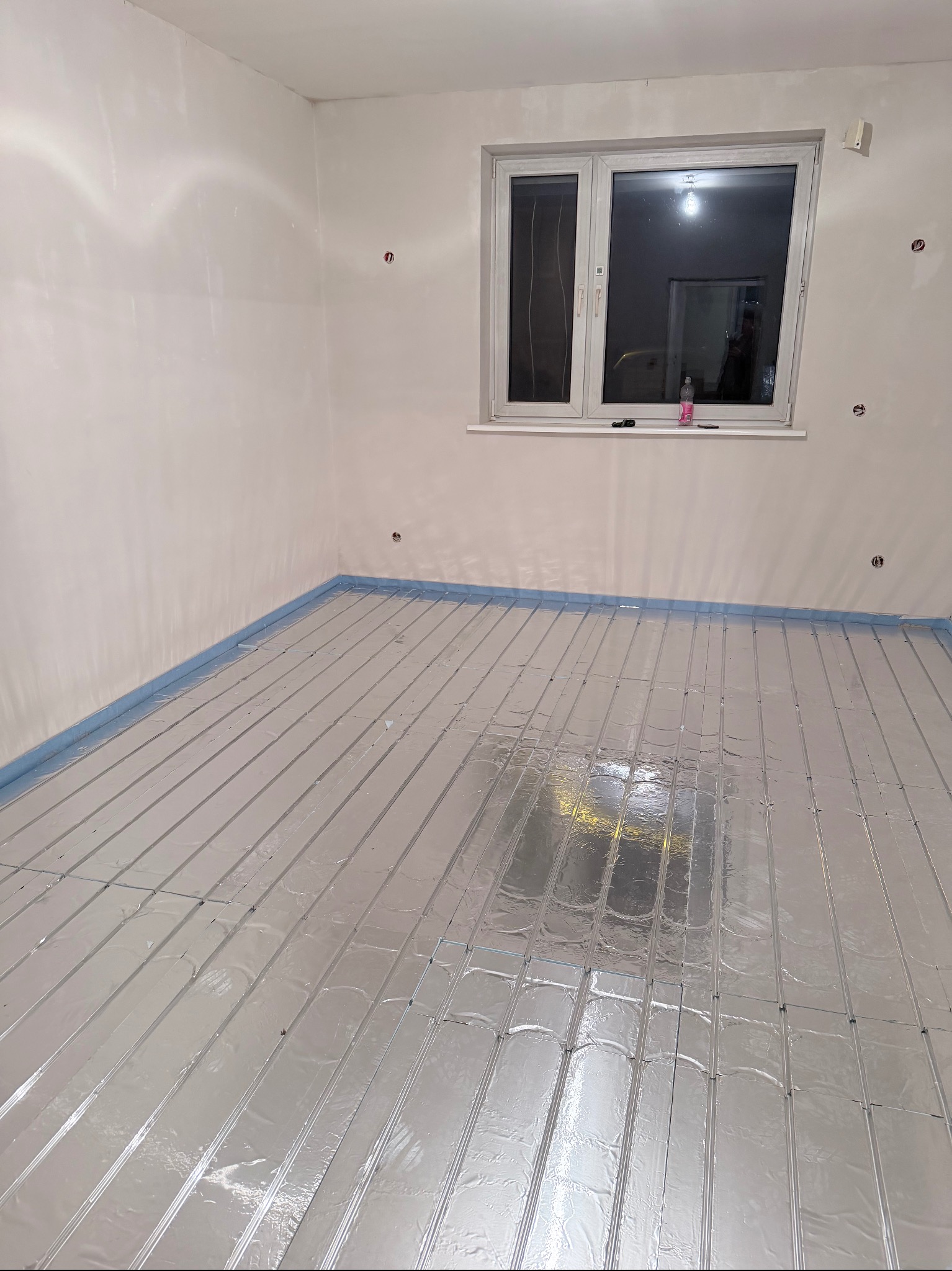 Ułożona instalacja ogrzewania podłogowego w pokoju z oknem, zabezpieczona niebieską folią przy ścianach. Widoczne otwory na gniazdka elektryczne.
