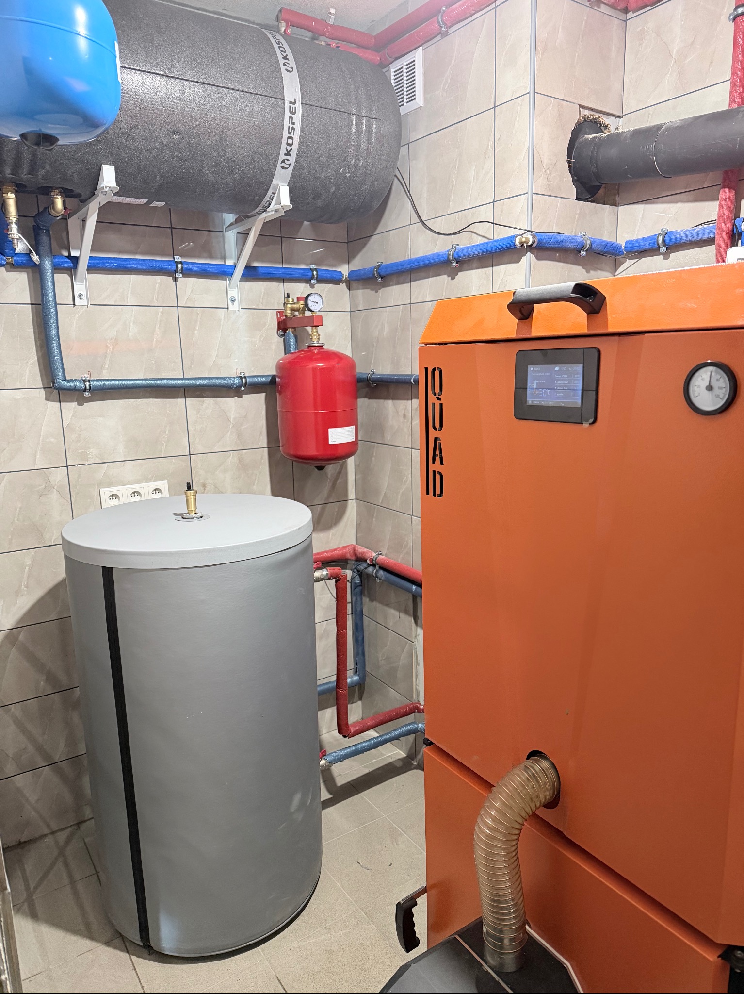 Pomarańczowy piec CO marki QUAD, szary zbiornik akumulacyjny i instalacja w kotłowni z beżowymi płytkami. Widoczne rury, zawory i manometr.