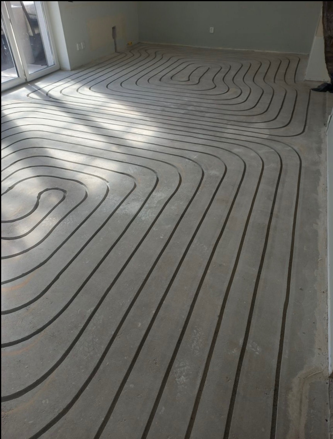 Ułożone rury ogrzewania podłogowego w surowym pomieszczeniu, widoczne złącza i fragment okna. Instalacja przed wylaniem posadzki, w jasnym świetle.