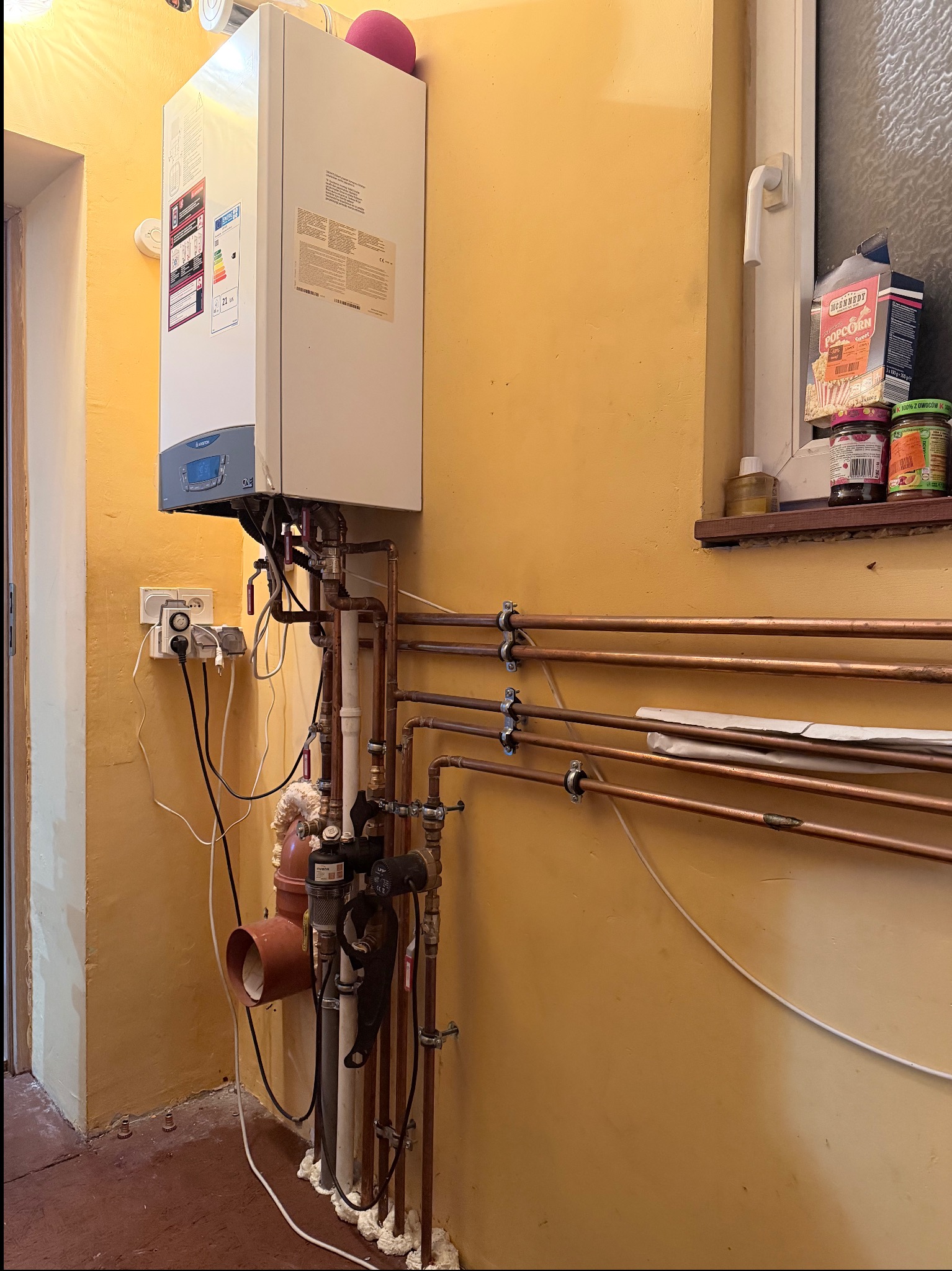 Biały piec gazowy Ariston ONE z miedzianą instalacją CO na żółtej ścianie, widok z boku. Na parapecie okna pudełko popcornu i słoiki. Widoczne gniazdka elektryczne.
