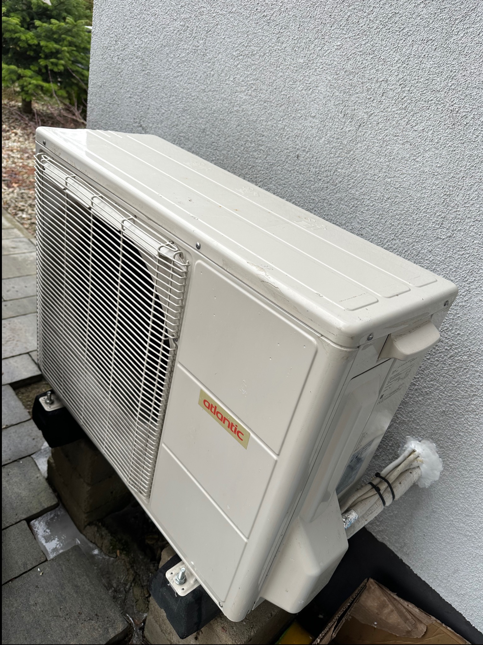 Klimatyzator Atlantic zamontowany na zewnątrz budynku, widoczne przewody i elementy mocujące. Ujęcie z dołu, podkreślające instalację urządzenia.