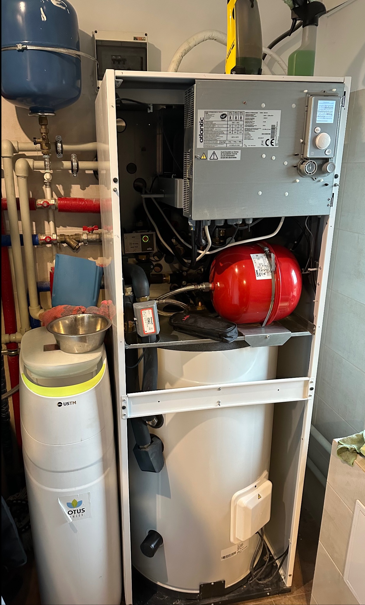 Biały piec grzewczy Atlantic z czerwonym zbiornikiem ekspansyjnym i filtrem wody Otus, widoczne rury instalacji CO w tle, narzędzia na półce wewnątrz pieca.