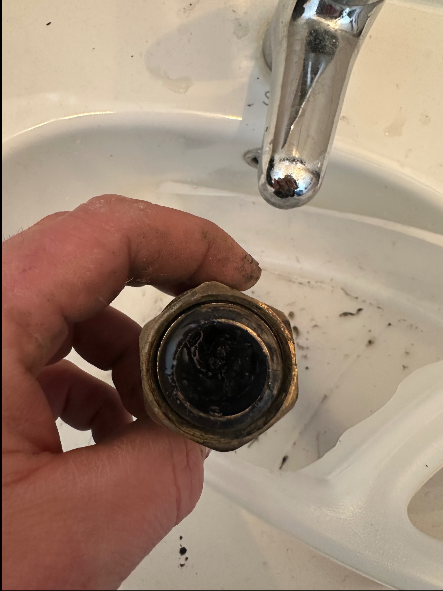 Zdemontowany, brudny zawór w dłoni nad umywalką z widocznym kranem. Detal instalacji hydraulicznej w trakcie naprawy lub wymiany.