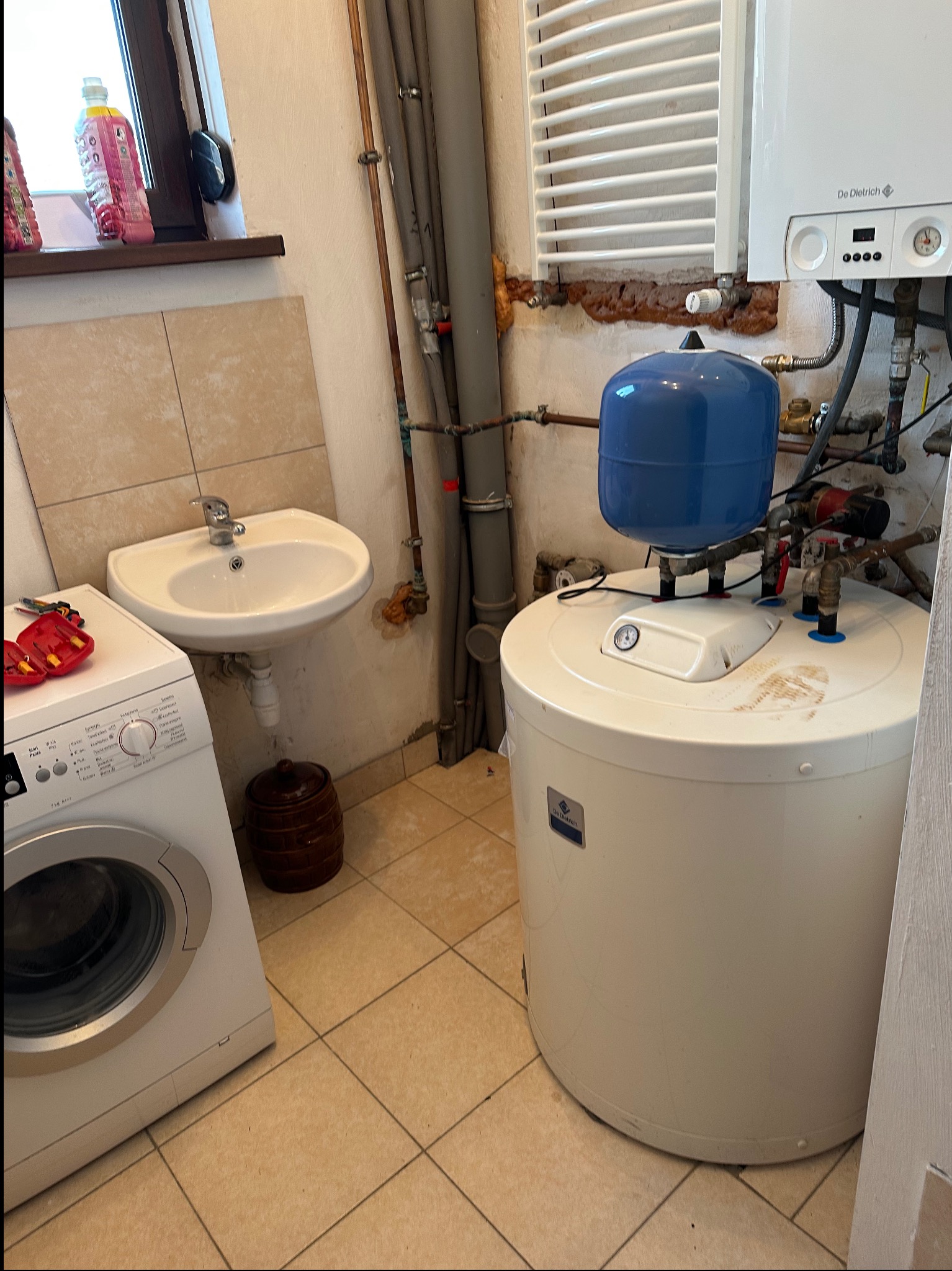 Biały bojler z niebieskim zbiornikiem wyrównawczym w pomieszczeniu gospodarczym, obok pralka i umywalka. Widoczne rury i grzejnik drabinkowy.
