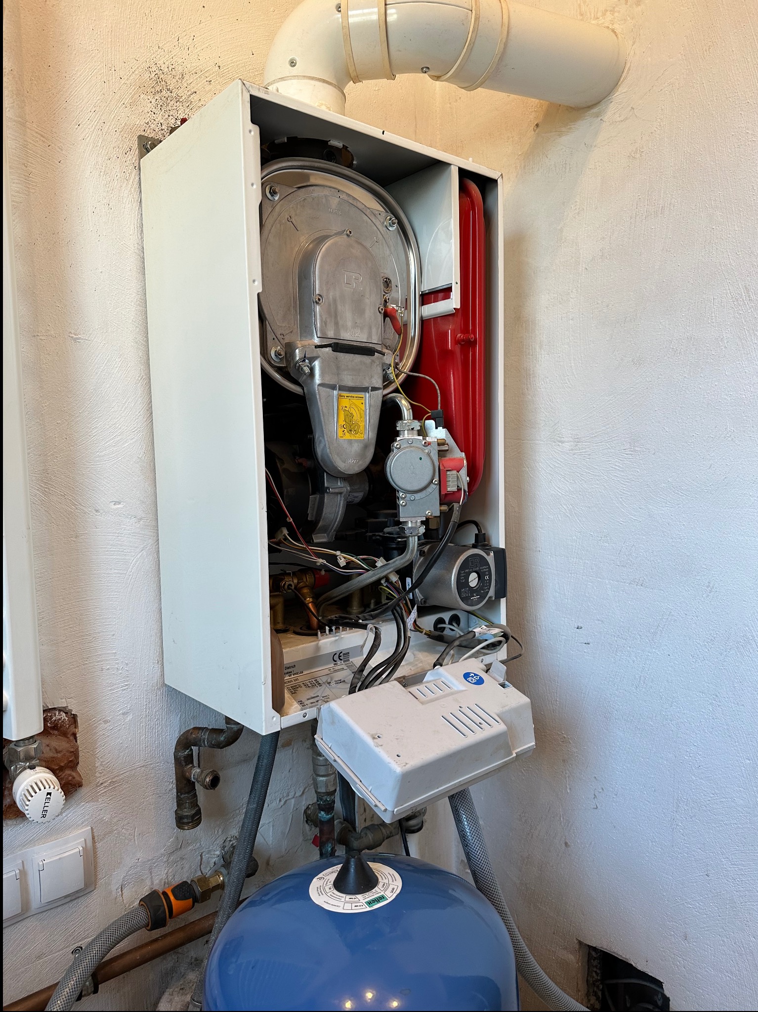 Otwarty, biały piec gazowy z widocznymi elementami wewnętrznymi, rurami i niebieskim zbiornikiem wyrównawczym na tle ściany o szorstkiej fakturze.