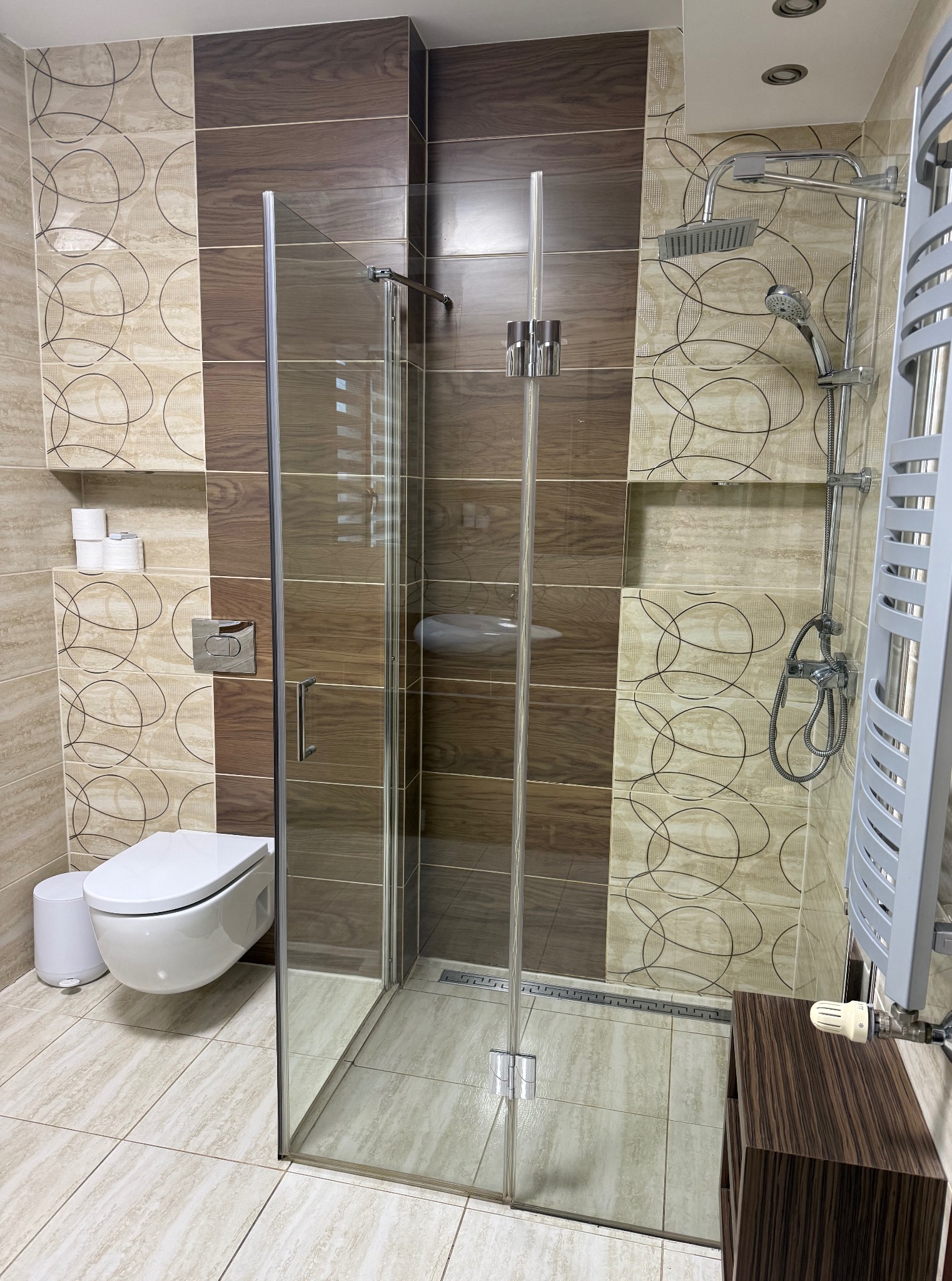 Nowoczesna łazienka z kabiną prysznicową typu walk-in, beżowo-brązowe płytki z motywem kół, podwieszana toaleta i grzejnik drabinkowy.