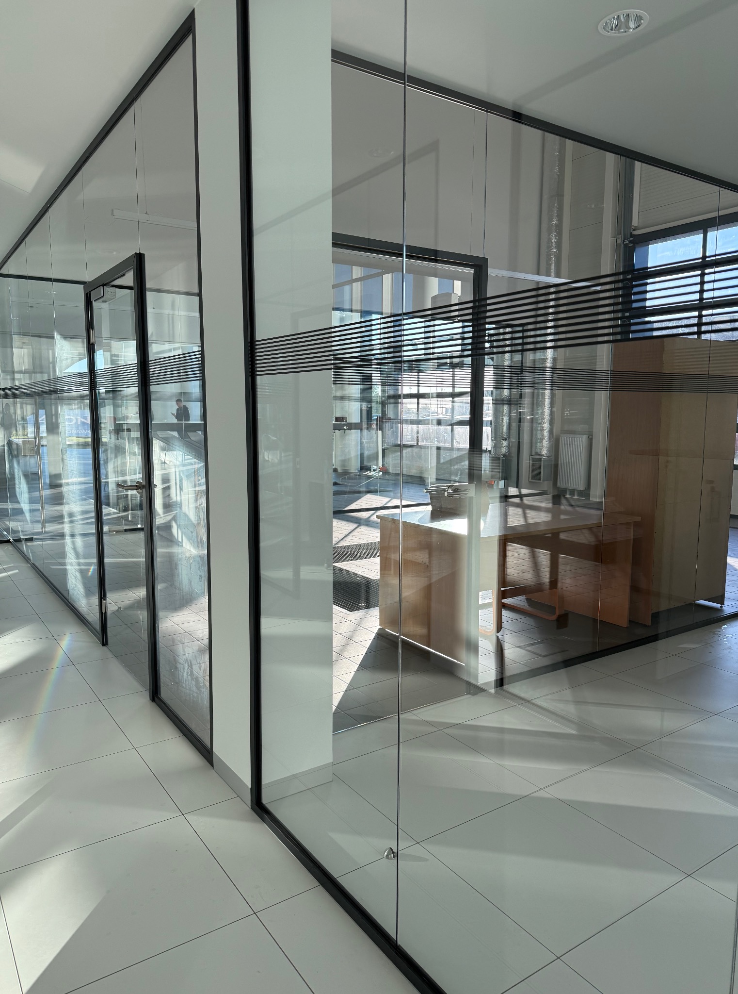 Nowoczesne biuro z przeszklonymi ścianami działowymi. Widoczny porządek i czystość, jasne płytki podłogowe i meble biurowe w tle. Minimalistyczny design.