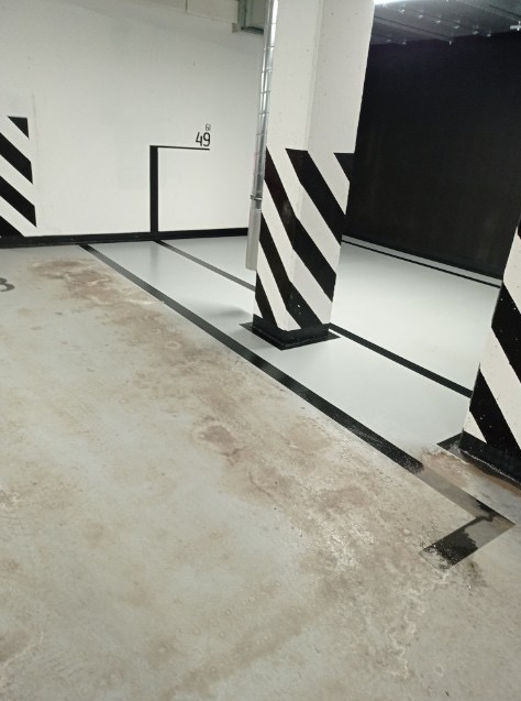 Brudna posadzka w garażu podziemnym z numerem stanowiska '49' na ścianie, czarno-białe pasy na filarach. Widoczne plamy i zabrudzenia na posadzce.