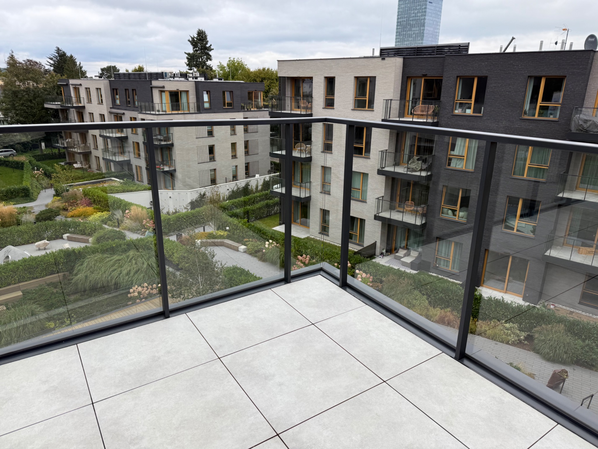 Nowoczesny balkon z szarymi płytkami i szklaną balustradą. Widok na zadbany ogród osiedlowy i budynki z balkonami. W tle wieżowiec.