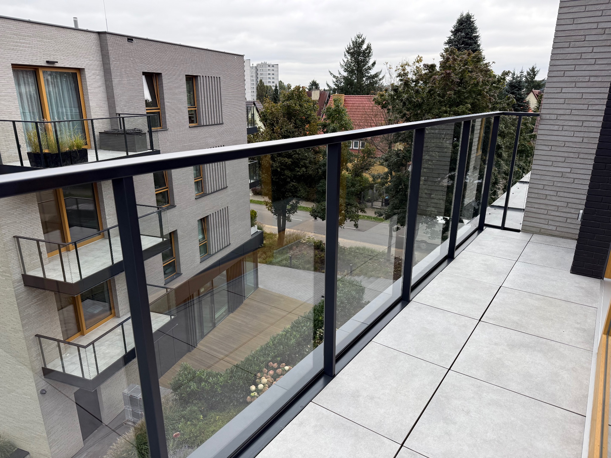 Nowoczesny balkon z szarymi płytkami i szklaną balustradą. Widok na zadbany dziedziniec z zielenią i architekturą miejską w tle. Minimalistyczny design, stonowana kolorystyka.