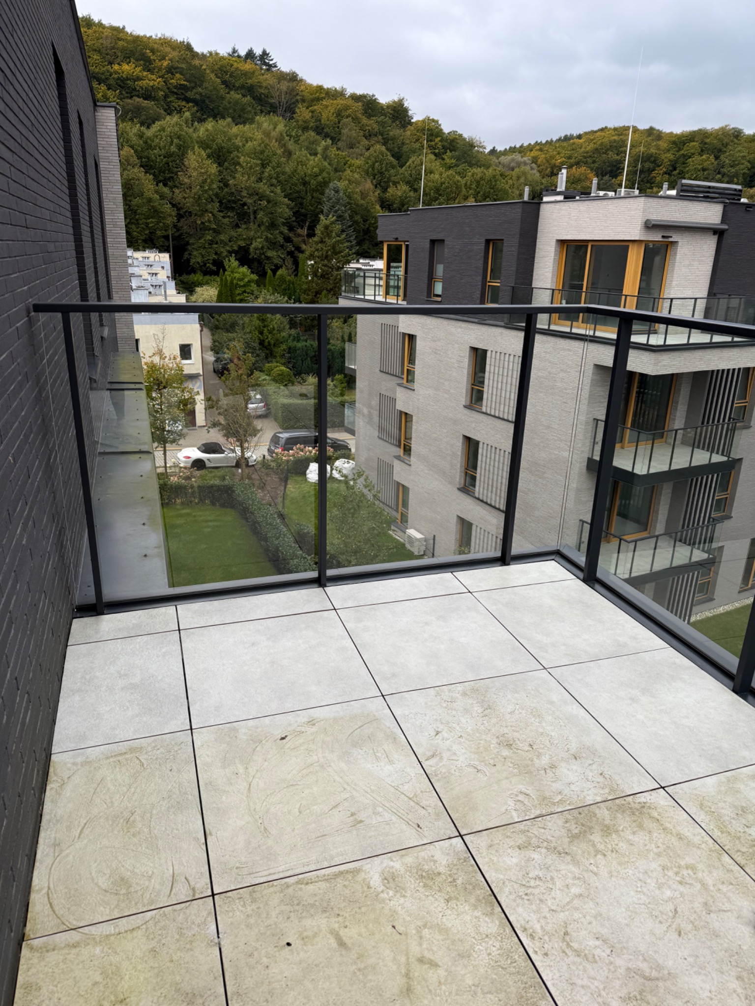 Szare płytki na balkonie apartamentu z widokiem na zieleń i budynki. Widoczne ślady użytkowania na płytkach, sugerujące potrzebę czyszczenia.