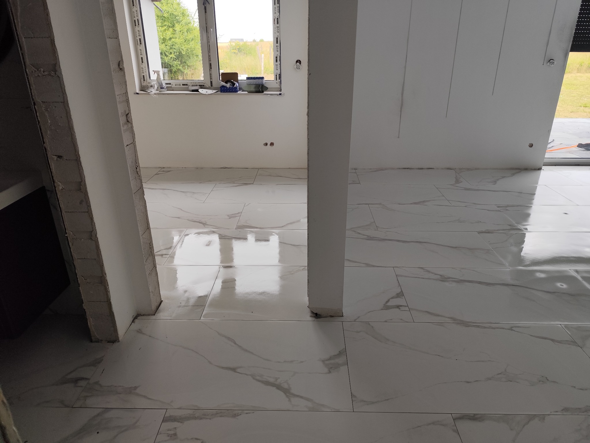 Wykończona podłoga z białych, marmurkowych płytek w nowym wnętrzu. Widoczne okno z akcesoriami i surowe wykończenie ościeżnicy.