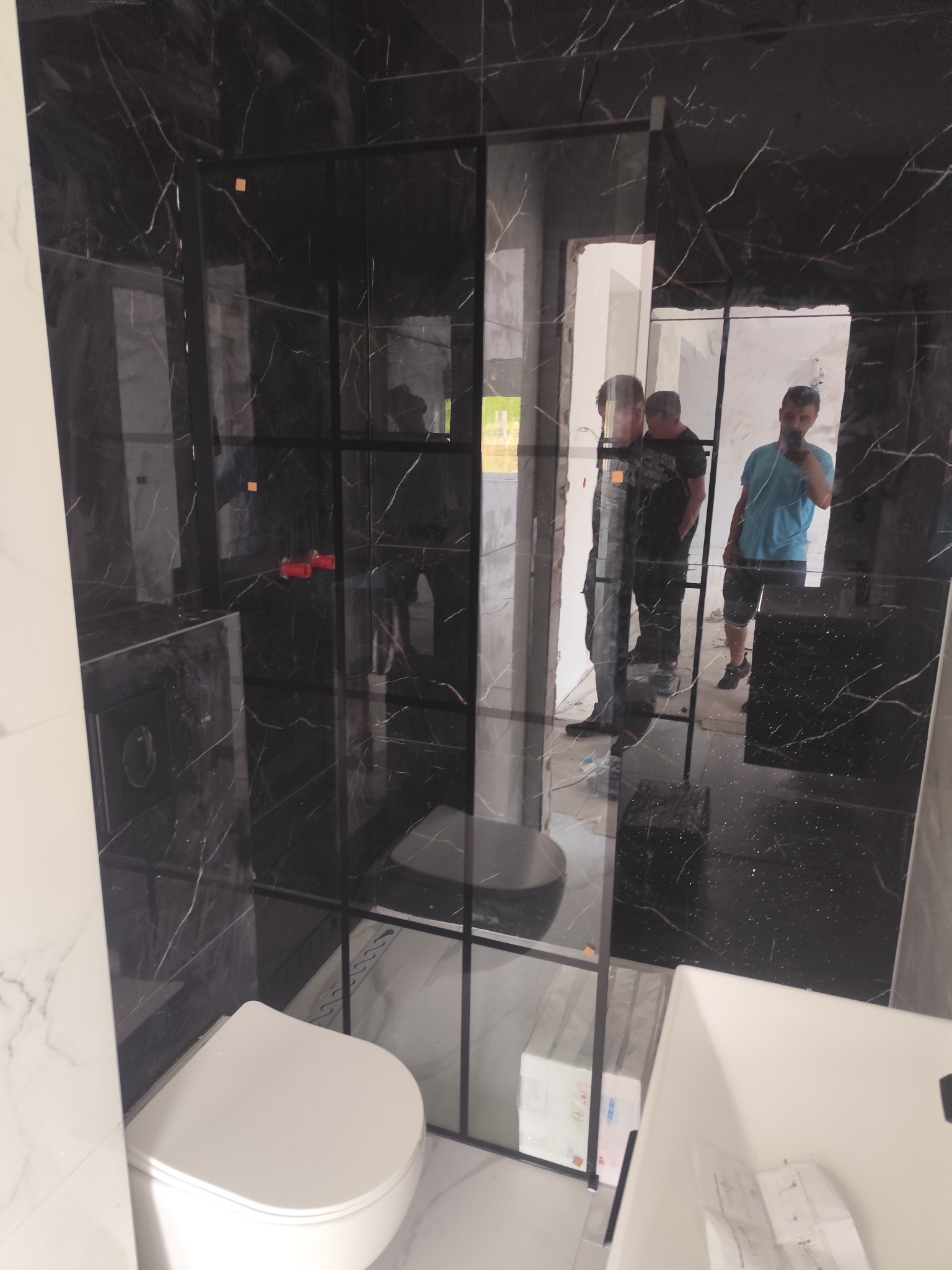 Wykończona łazienka z czarnymi płytkami imitującymi marmur, białą armaturą i kabiną prysznicową z czarnymi profilami. W tle odbicie osób w lustrze.