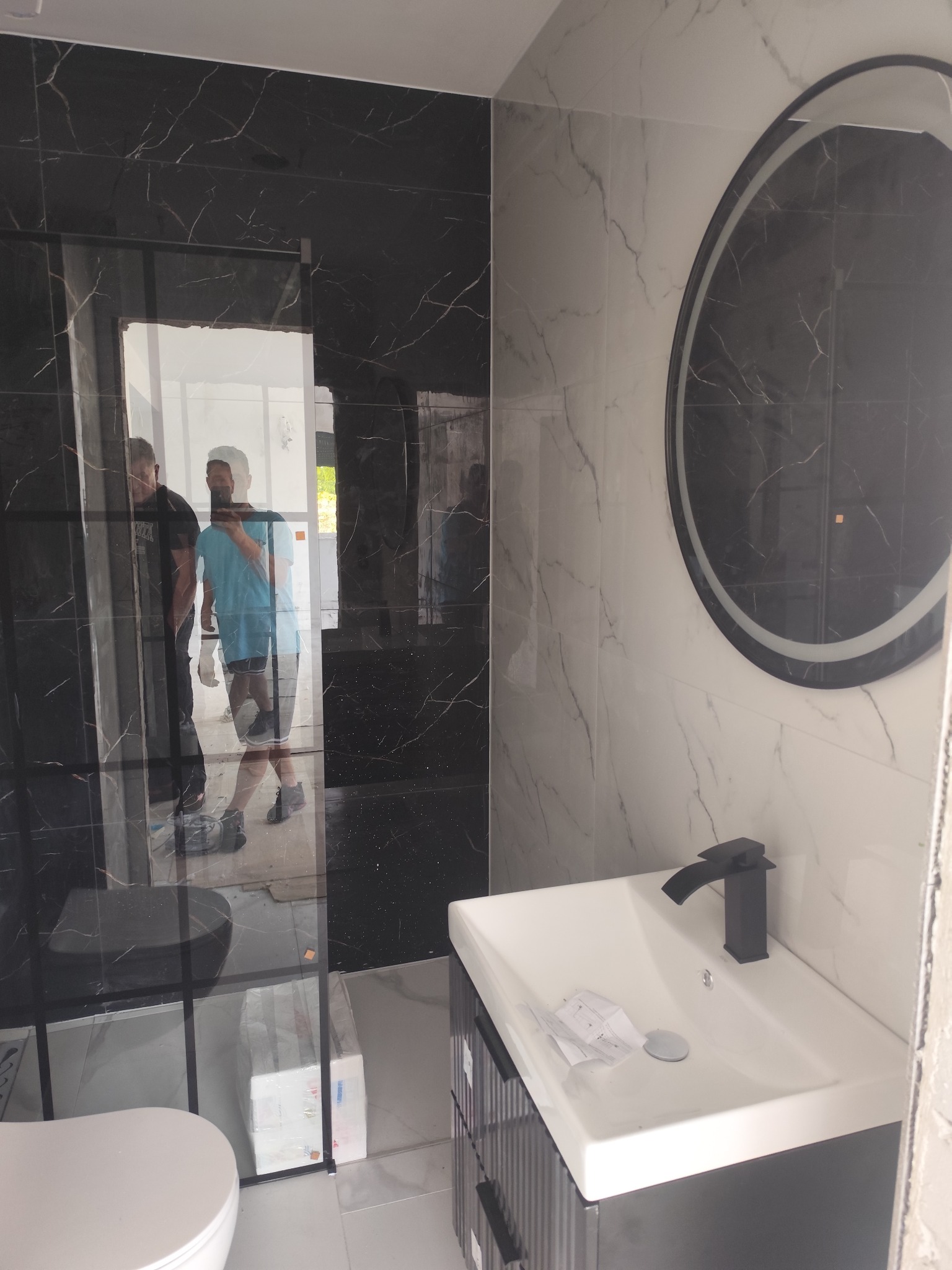 Wykończona łazienka z czarno-białymi płytkami imitującymi marmur, okrągłym lustrem z podświetleniem i czarną armaturą, widok na kabinę prysznicową.