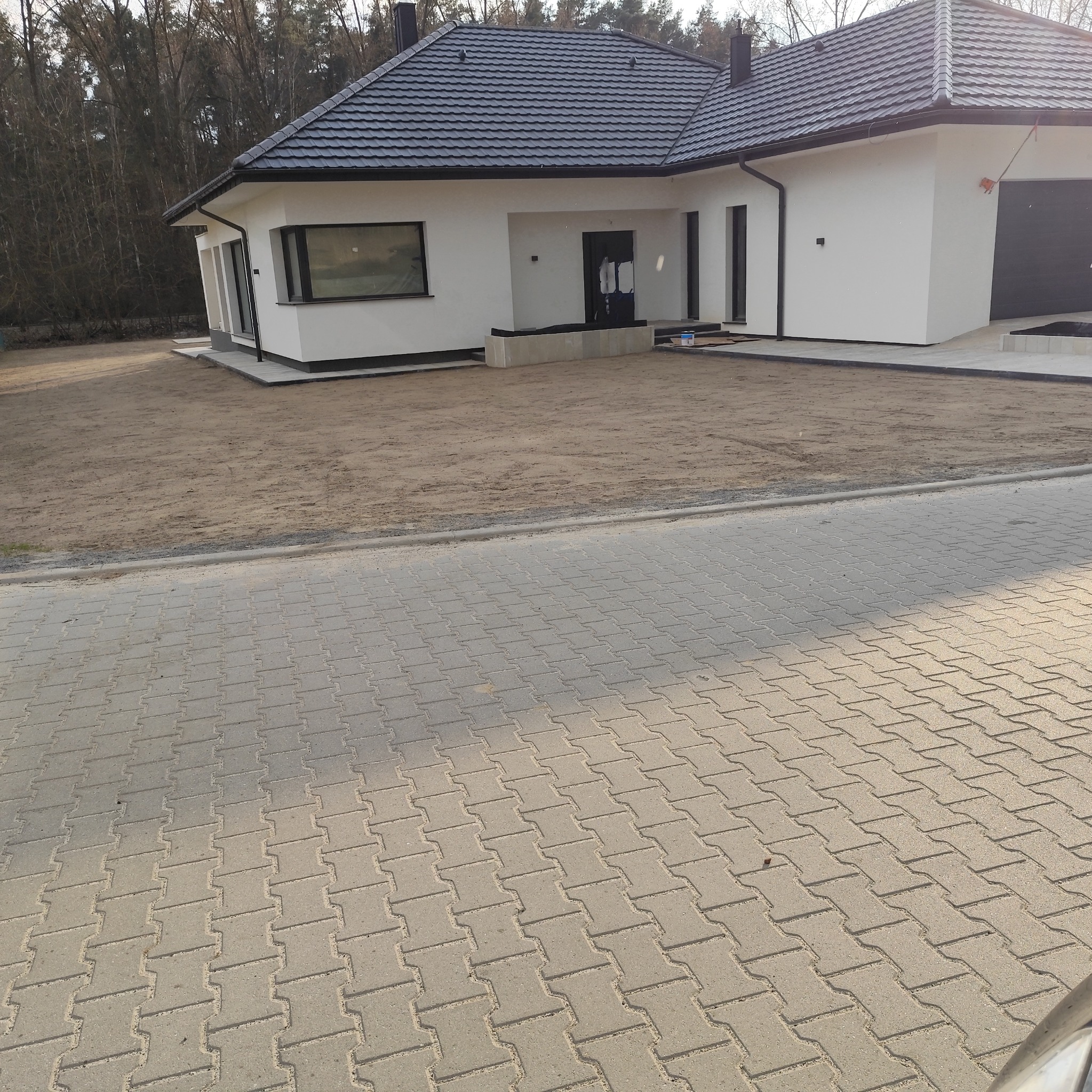 Nowoczesny dom jednorodzinny z czarnym dachem i białymi ścianami, widok z poziomu chodnika, teren przygotowany pod trawnik, drzewa w tle.