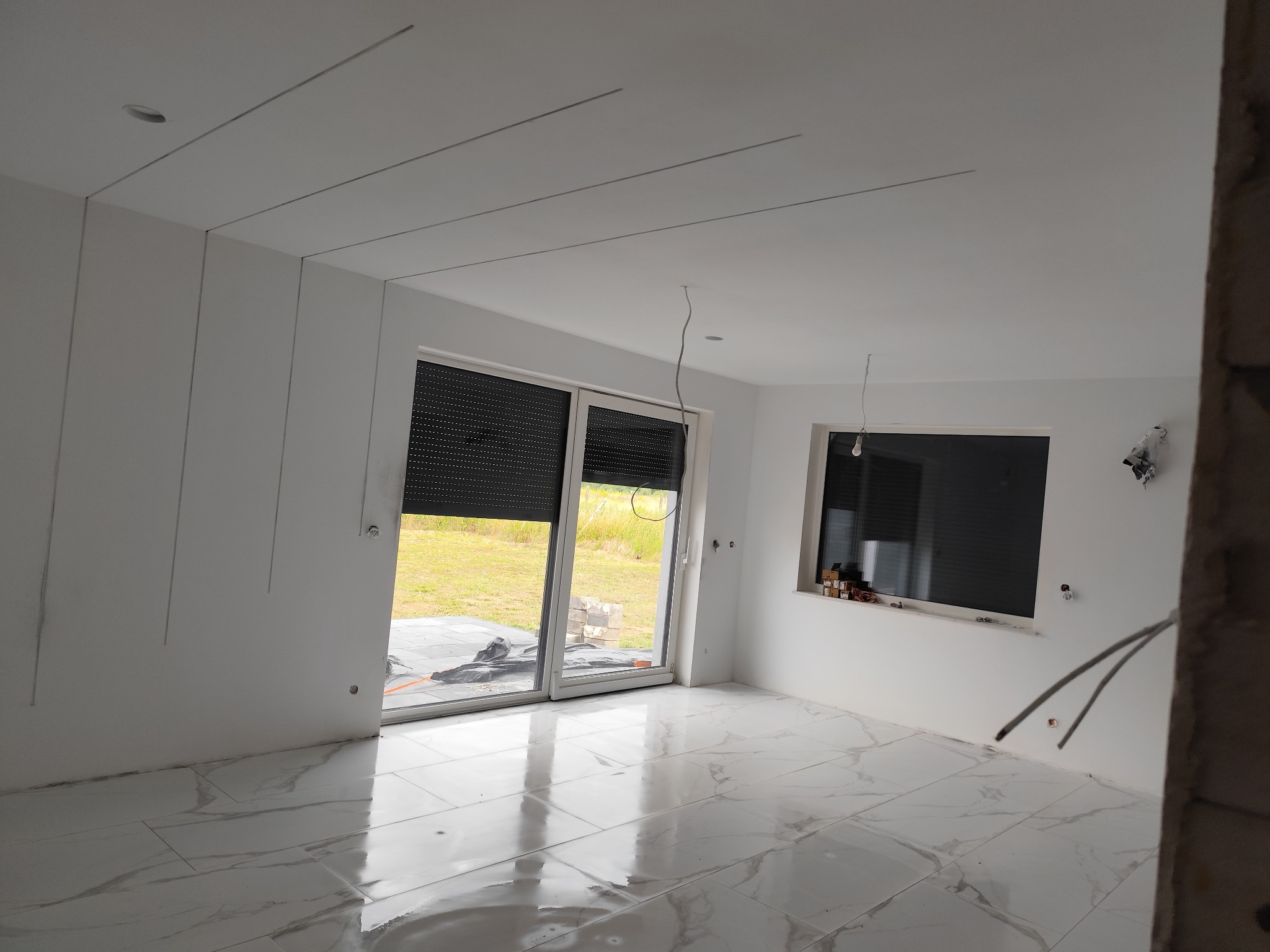 Jasne wnętrze w trakcie wykańczania: białe ściany z listwami, marmurowe płytki podłogowe, okna z roletami. Widoczne przewody elektryczne i niedokończone detale.