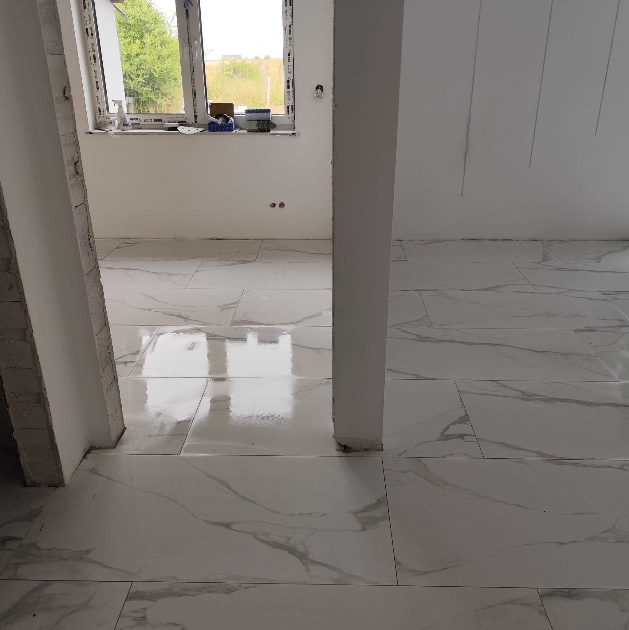 Białe, błyszczące płytki imitujące marmur ułożone na podłodze w nowym, jasnym wnętrzu. Widoczny fragment okna i surowa kolumna.