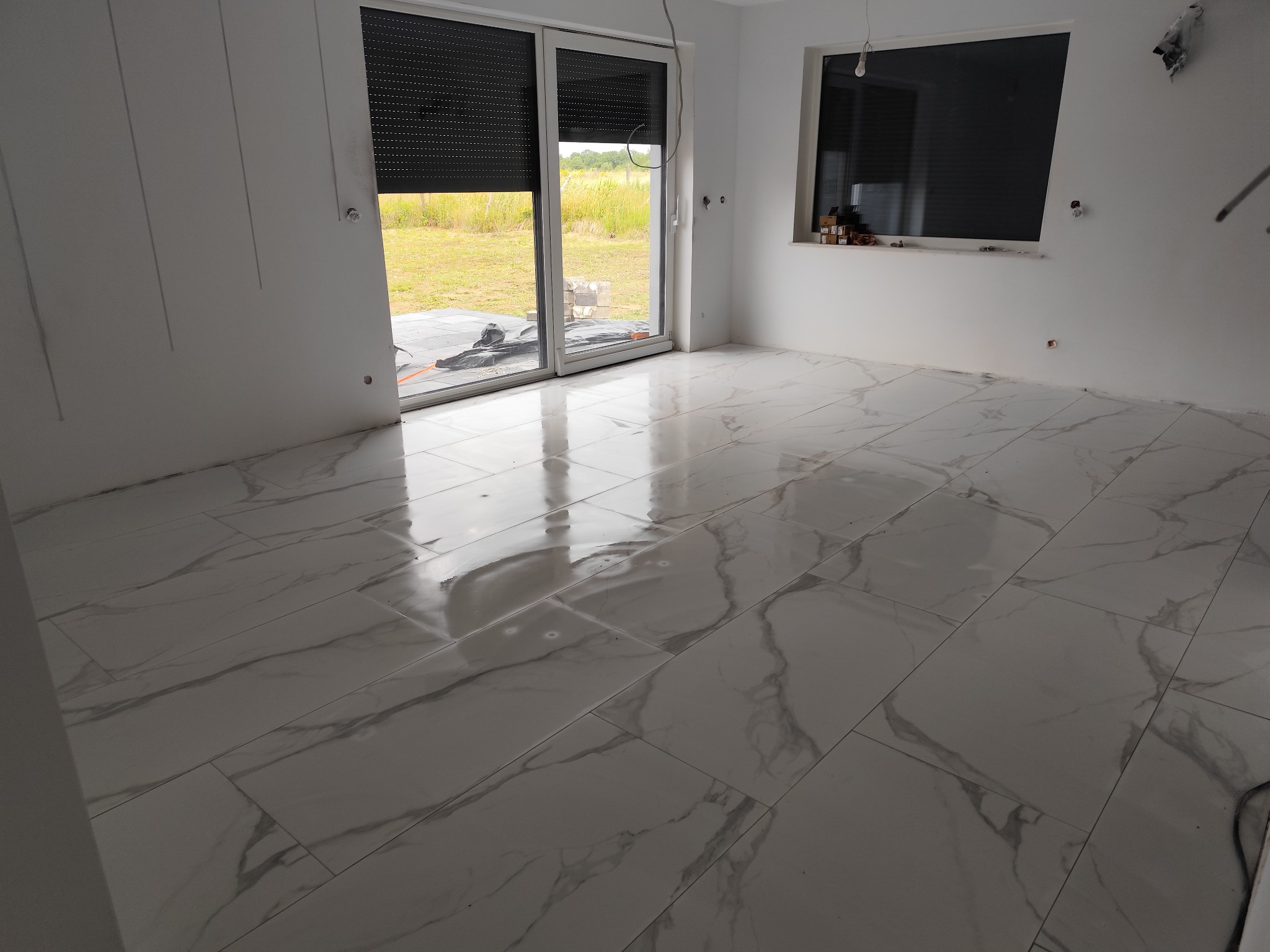 Jasne, duże płytki imitujące marmur na podłodze w nowym pomieszczeniu z oknami i roletami. Widoczne ślady po pracach wykończeniowych.