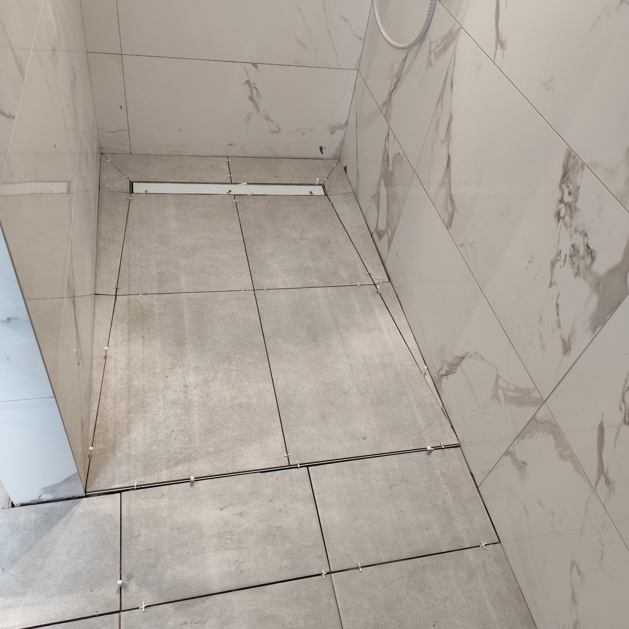 Wykończony brodzik z odpływem liniowym, szare płytki podłogowe i białe marmurowe płytki ścienne. Widoczne krzyżyki dystansowe. Minimalistyczny design.