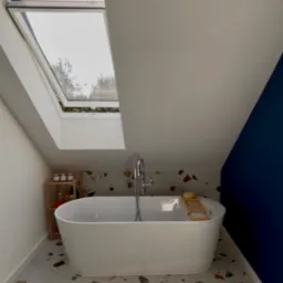 Biała wanna wolnostojąca w łazience na poddaszu z oknem dachowym, mozaikowa podłoga i niebieska ściana. Drewniana półka z kosmetykami obok wanny.