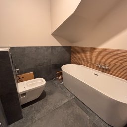 Modern Impact - Nowoczesna łazienka w trakcie montażu: biała wanna wolnostojąca z drewnianym panelem ściennym, podwieszana toaleta z kartonem obok, szare płytki podłogowe i ścienne.