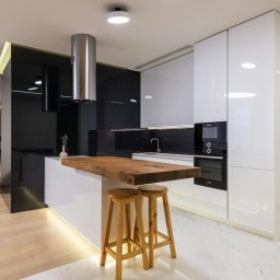 Modern Impact - Nowoczesna kuchnia z wyspą: białe szafki, drewniany blat, stalowy okap. Minimalistyczny design, oświetlenie LED pod wyspą, marmurowe płytki podłogowe.