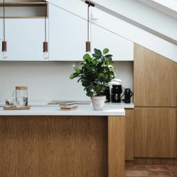 Modern Impact - Jasna kuchnia na poddaszu z drewnianą wyspą, rośliną w doniczce i trzema lampami wiszącymi. Uwagę przykuwa terakotowa podłoga i skośny sufit.