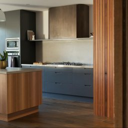 Modern Impact - Nowoczesna kuchnia z drewnianą wyspą, szarymi szafkami i betonowym blatem. Minimalistyczny design z rośliną w złotej doniczce i drewnianą ścianką działową.