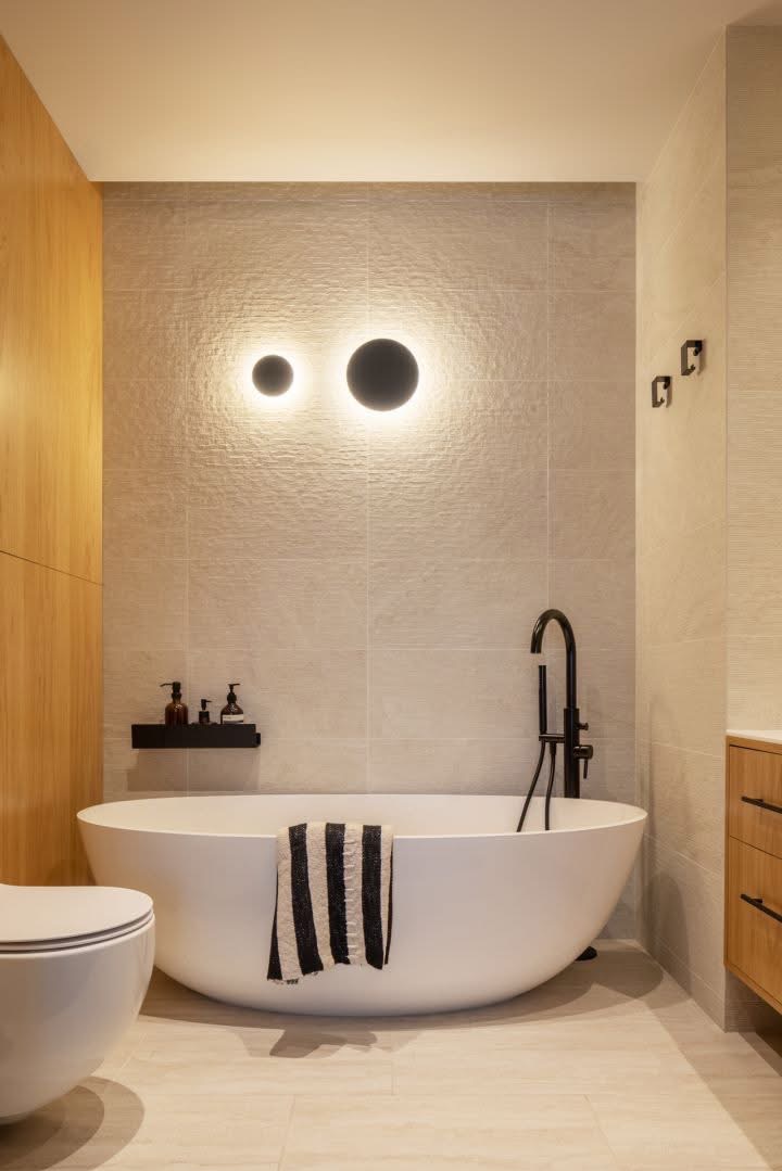 Nowoczesna łazienka z wolnostojącą wanną, czarną armaturą i okrągłymi lampami ściennymi. Beżowe płytki z delikatną fakturą. Minimalistyczny design.