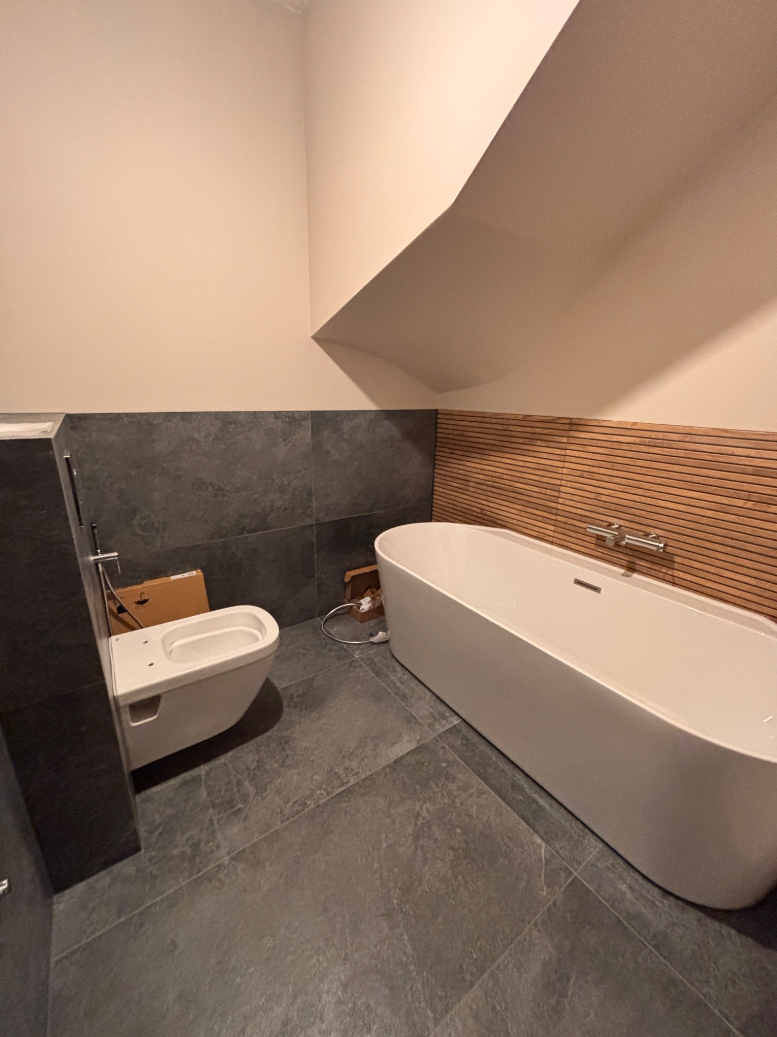 Nowoczesna łazienka w trakcie montażu: biała wanna wolnostojąca z drewnianym panelem ściennym, podwieszana toaleta z kartonem obok, szare płytki podłogowe i ścienne.